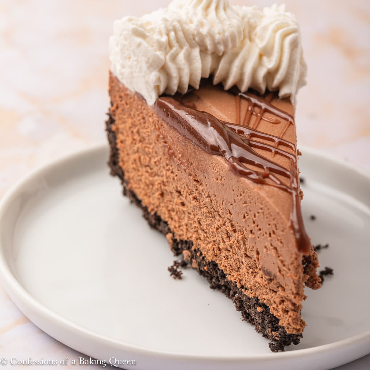 A slice of chocolate cheesecake with an Oreo crust, topped with chocolate ganache and whipped cream, served on a white plate.