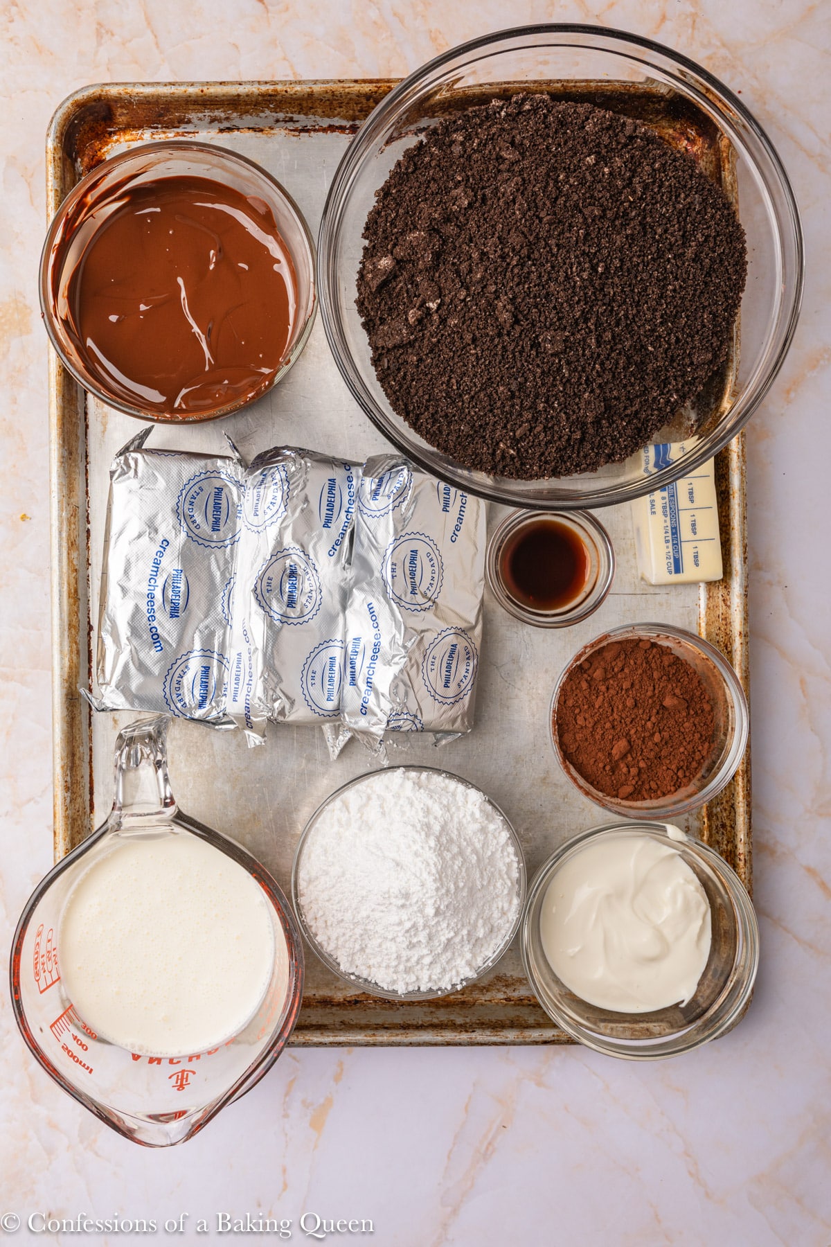 A baking tray holds bowls and containers with ingredients: chocolate, crushed cookies, cocoa powder, vanilla, cream, butter, powdered sugar, cream cheese, and sour cream, all arranged neatly from a top-down view.
