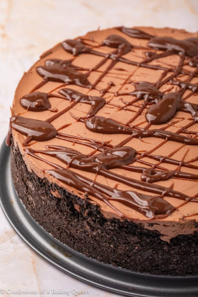 A close-up of a chocolate cheesecake with a thick Oreo cookie crust, topped with a smooth chocolate layer and drizzled with glossy chocolate sauce, presented on a round metal pan.