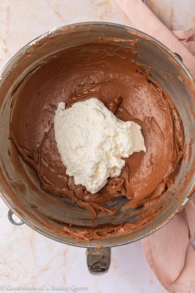 A mixing bowl containing chocolate batter with a large dollop of white ricotta cheese in the center, sitting on a marble surface next to a pink cloth.