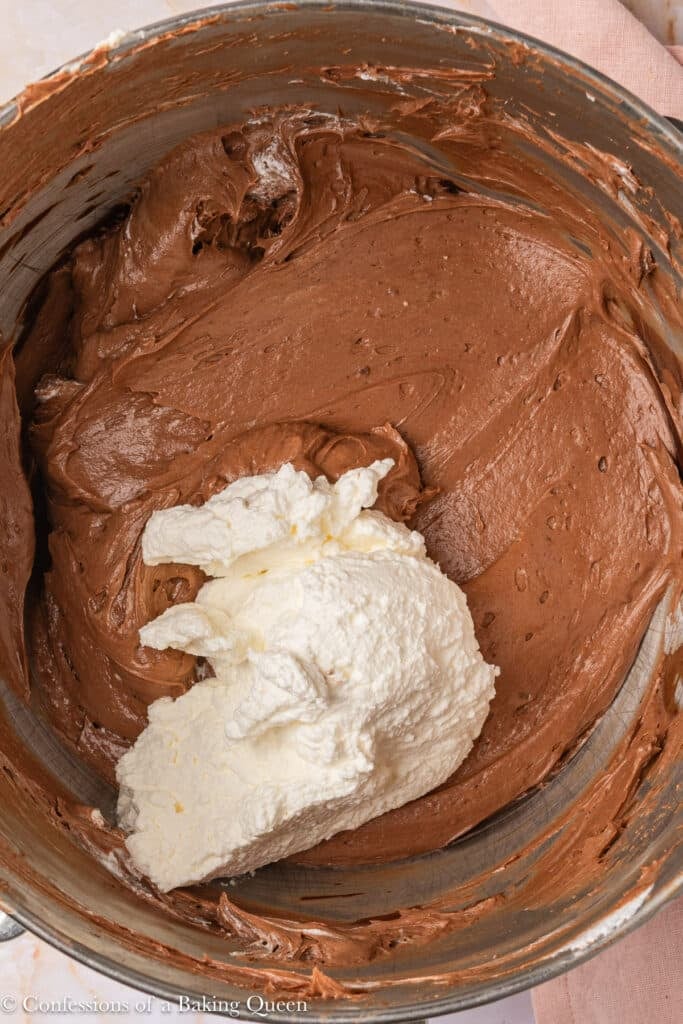A mixing bowl filled with chocolate batter, with a large dollop of whipped cream or frosting sitting on top, ready to be folded in.