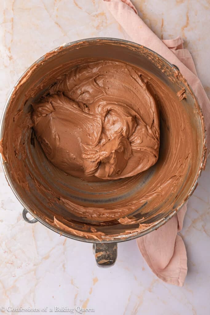 A metal mixing bowl filled with creamy chocolate frosting sits on a marble surface, with a light pink cloth partially visible beside it.