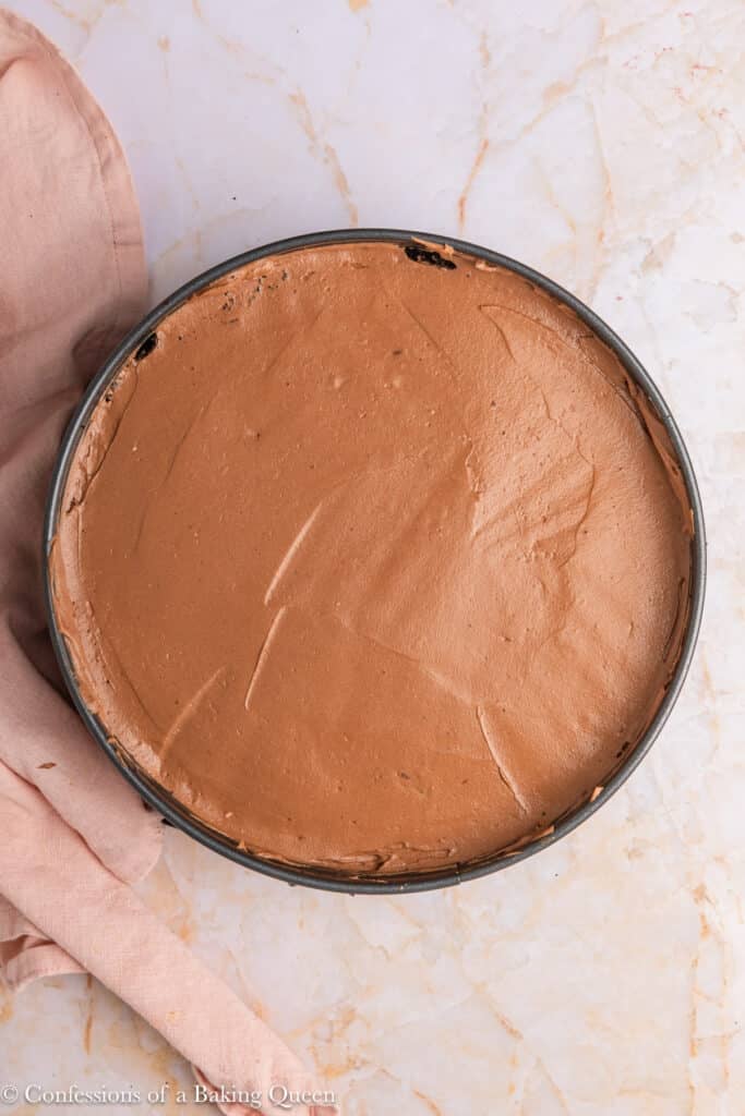 A round chocolate cheesecake sits in a springform pan on a light marble surface, with a pink cloth partially visible to the left. The cheesecake has a smooth, creamy top with a few visible marks.