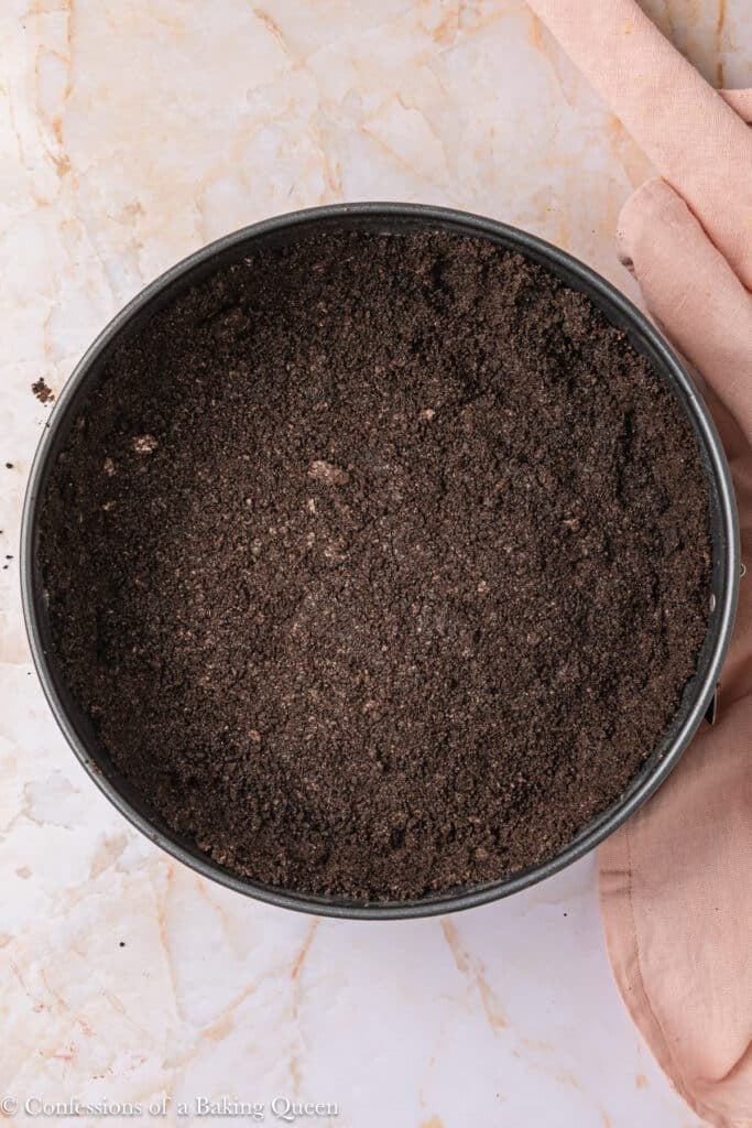 A round pan filled with a pressed chocolate cookie crumb crust sits on a marble surface next to a light pink cloth.