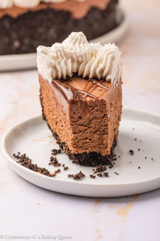 A slice of chocolate cheesecake with a layer of chocolate ganache, whipped cream topping, and a chocolate cookie crust sits on a white plate. Crumbs are scattered around the slice.
