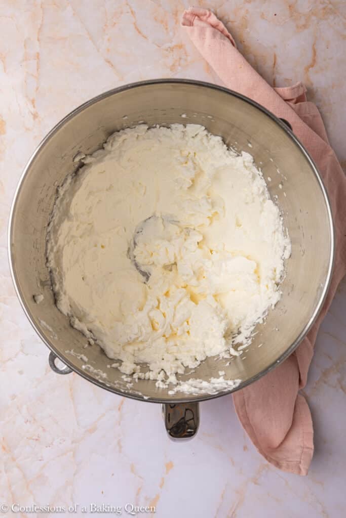 A metal mixing bowl filled with whipped cream cheese mixture sits on a marble countertop next to a pink cloth napkin.