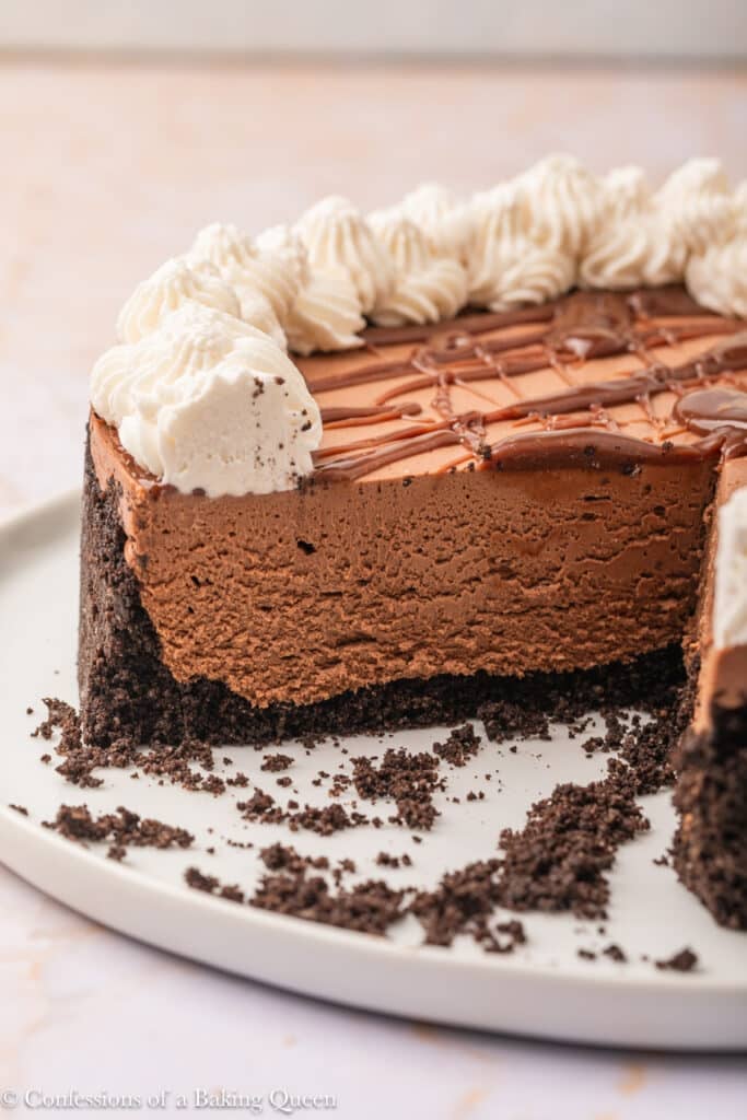 A rich, creamy chocolate cheesecake with a chocolate cookie crust is sliced, revealing its smooth texture. The cake is topped with piped whipped cream and a drizzle of chocolate, with crumbs scattered on a white plate.