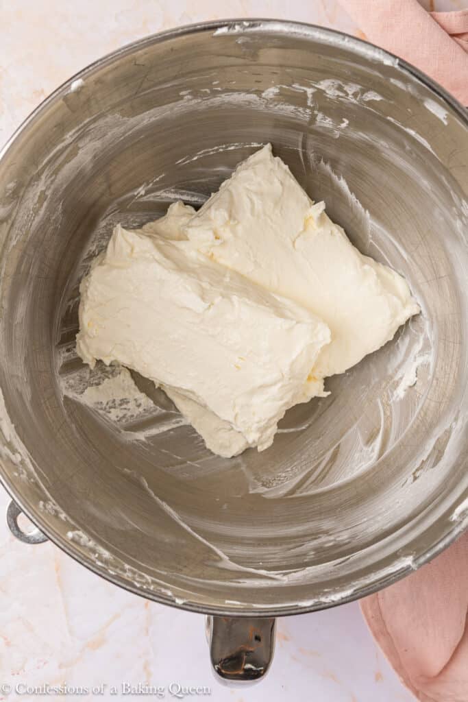 A metal mixing bowl containing two blocks of softened cream cheese, partially blended, with a creamy texture visible against the bowl's sides.