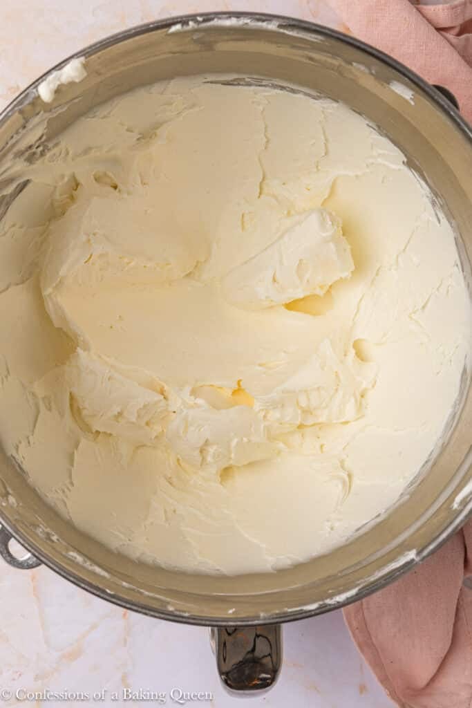 A metal mixing bowl filled with smooth, creamy, pale yellow buttercream frosting sits on a light-colored surface with part of a pink cloth visible at the edge.