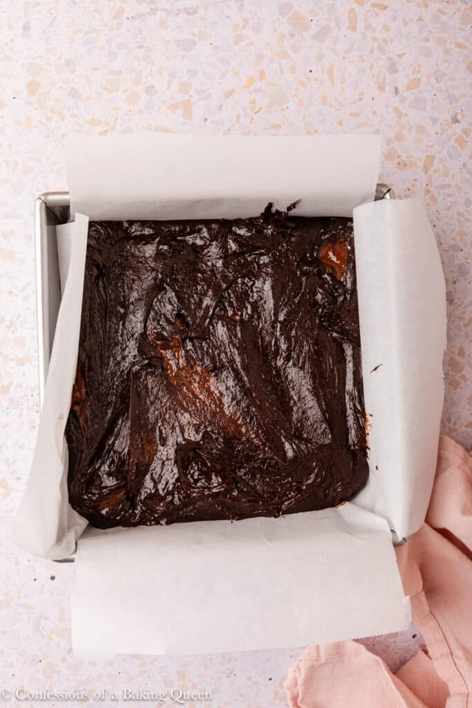 A square baking pan lined with parchment paper, filled with unbaked dark chocolate brownies batter, sits on a light countertop next to a pink cloth.