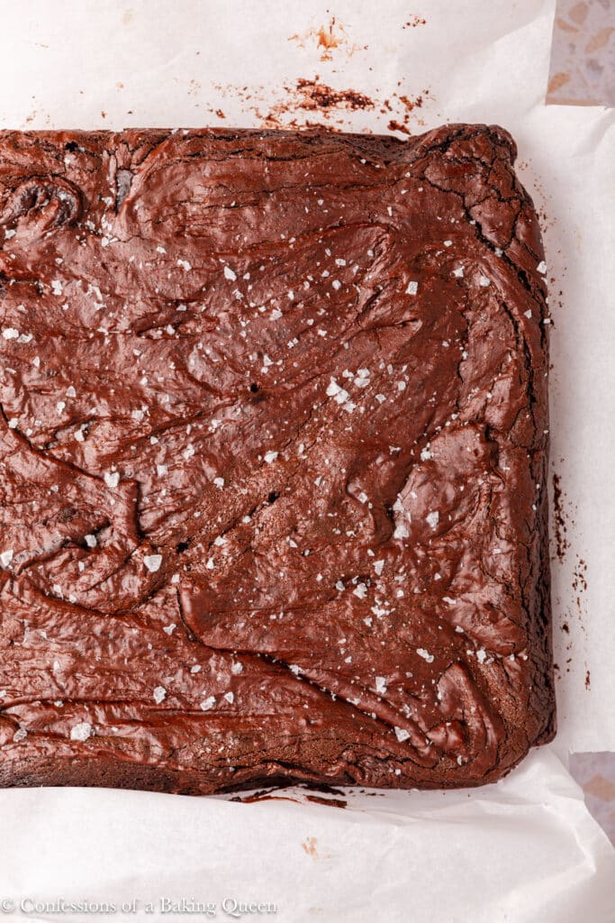 A close-up of a large, freshly baked pan of rich, fudgy dark chocolate brownies topped with flaky sea salt, resting on white parchment paper.