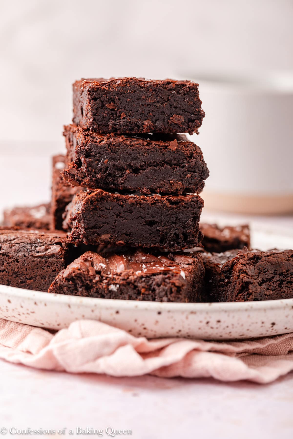 A stack of rich, fudgy dark chocolate brownies sits on a speckled plate, with a pink cloth underneath and a blurred mug in the background. The brownies have a glossy top and appear dense and moist.