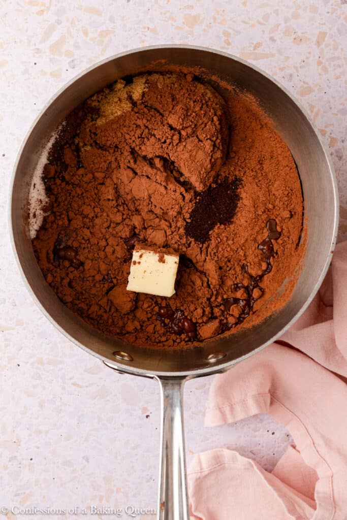 A saucepan containing cocoa powder, brown sugar, chocolate chips for dark chocolate brownies, a pat of butter, and instant coffee granules sits on a light-colored countertop with a pink cloth beside it.