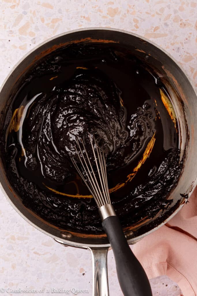 A saucepan filled with glossy, thick chocolate for dark chocolate brownies is being mixed with a black-handled whisk, resting on a light speckled countertop with a pink cloth nearby.