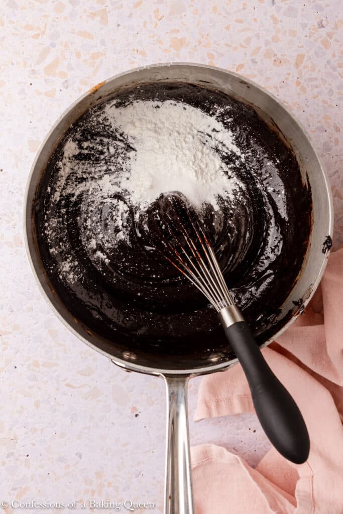 A whisk rests in a saucepan with dark chocolate brownie batter, partially mixed with white flour on top. The saucepan sits on a light speckled surface, next to a light pink cloth.