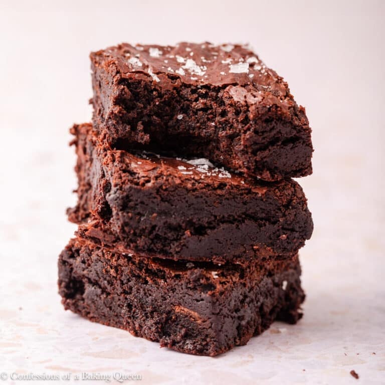 A stack of three rich, fudgy dark chocolate brownies with a sprinkling of flaky sea salt on top, set on a light-colored surface. The top brownie has a bite taken out of it.
